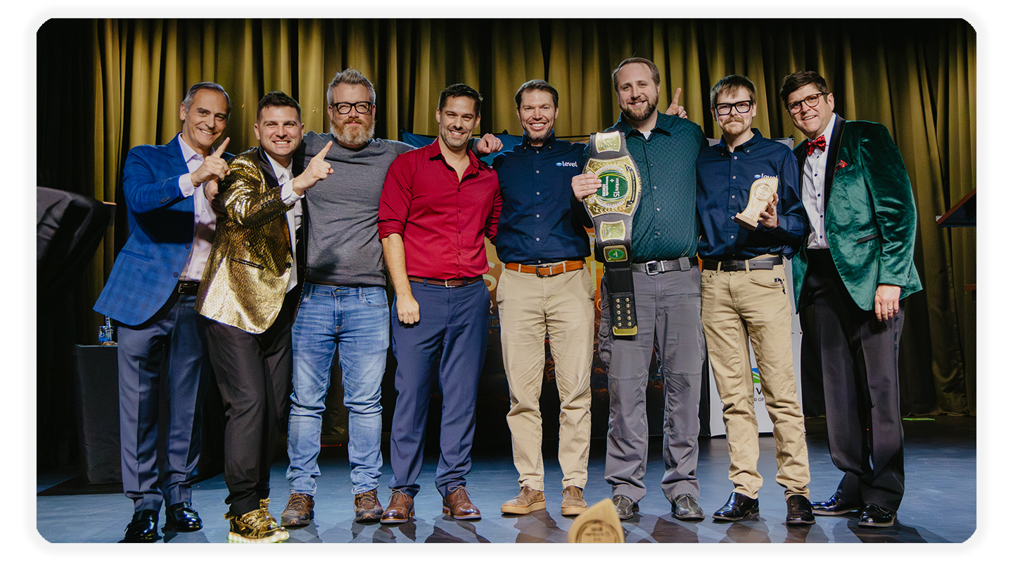 A group of eight people posing together on a stage, with two holding championship-style belts.